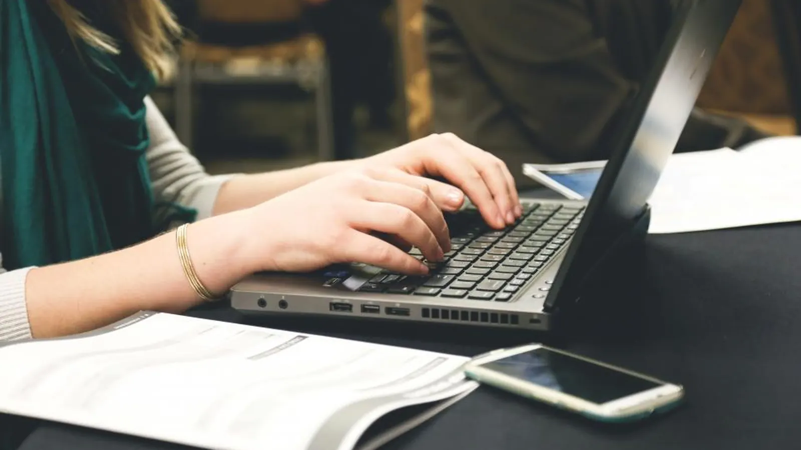 Hands typing on a laptop next to a smartphone and notebook